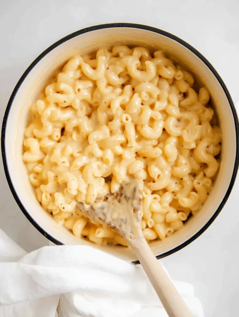 An overhead view of creamy mac and cheese in a white pot with a black rim, a wooden spoon inside, on a bright white surface.