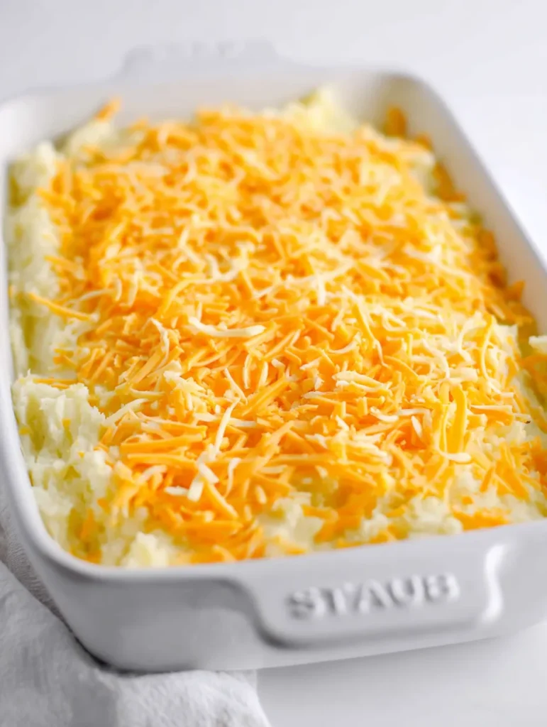 A white rectangular casserole dish filled with creamy mashed potatoes topped with shredded orange and white cheese.
