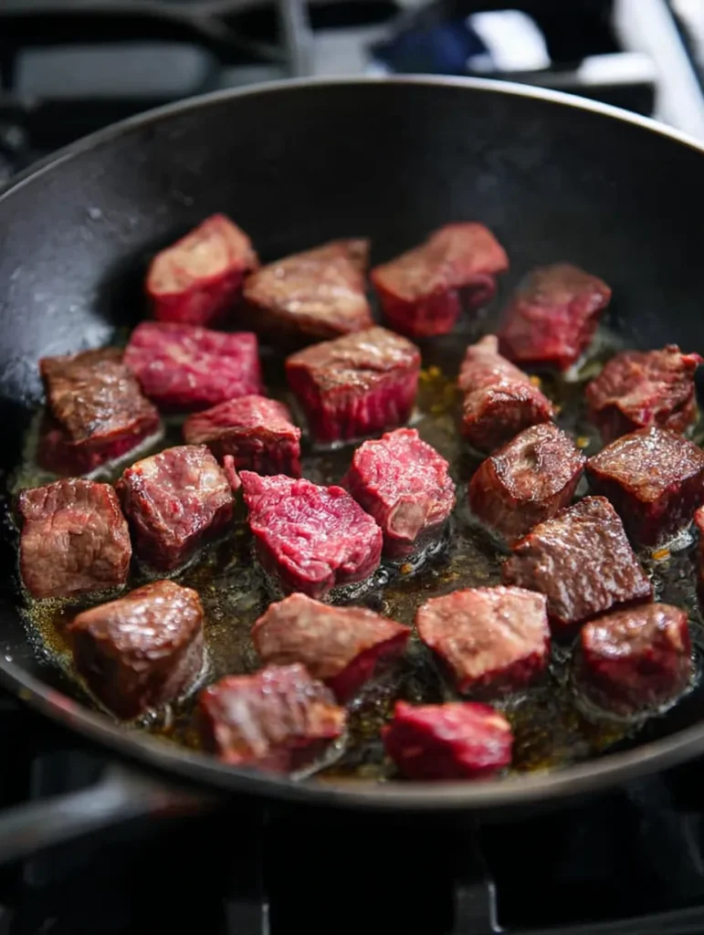 Close-up of beef cubes searing in a dark cast iron pan on a black gas stove, with some meat browned and others pink.